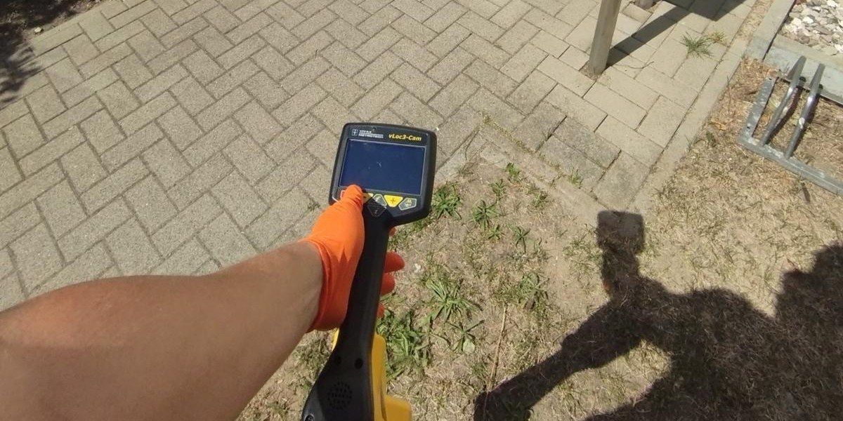 Mitarbeiter hält einen Ortungsempfänger über gepflastertem Boden vor einem Hauseingang