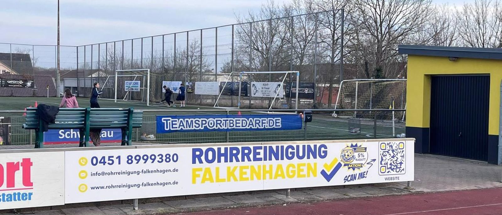 Bandenwerbung der Rohrreinigung Falkenhagen am Rand eines Sportplatzes mit Kunstrasen und Laufbahn