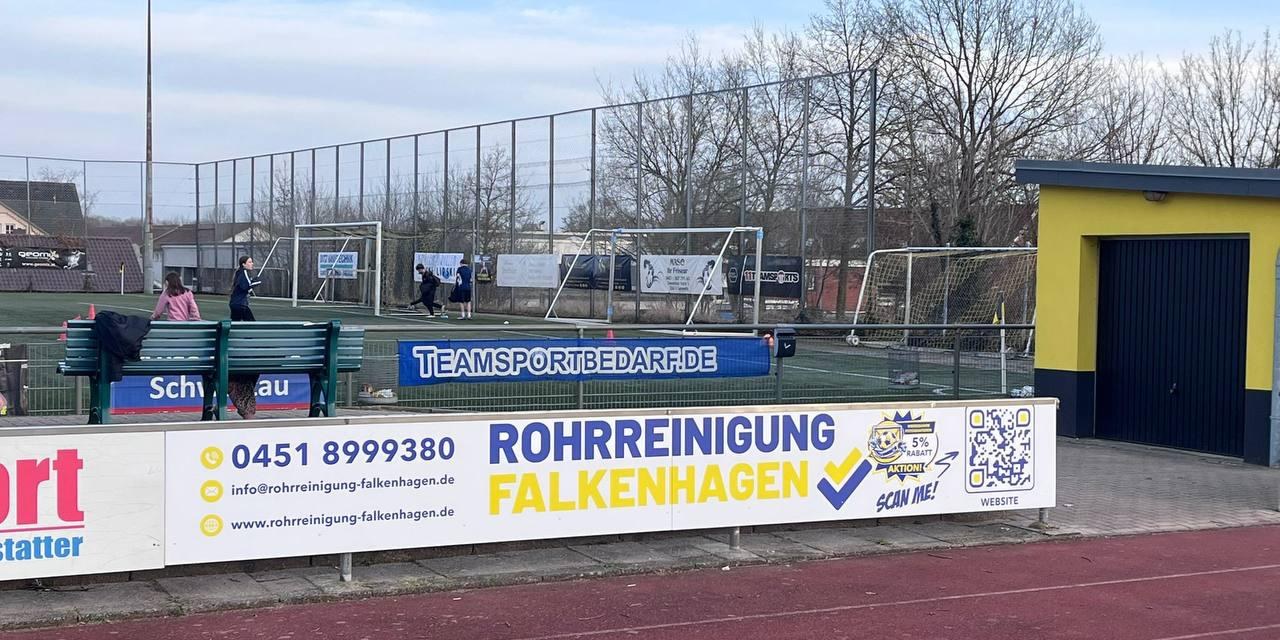 Bandenwerbung der Rohrreinigung Falkenhagen am Rand eines Sportplatzes mit Kunstrasen und Laufbahn